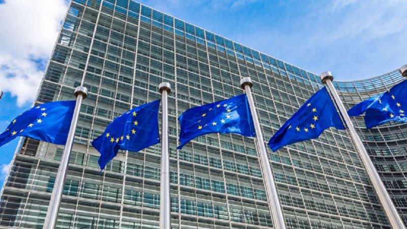 6 EU-Flaggen an Fahnenmasten vor einem mehrstöckigen Glasbau.