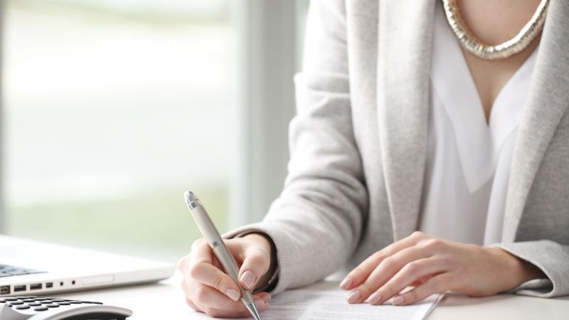 Das Bild zeigt einen Schreibtisch mit Laptop und Taschenrechner, an dem eine Frau ein Formular mit einem Stift ausfüllt.