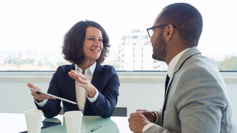 Das Bild zeigt zwei Menschen im Anzug, die sich anlächeln, während sie etwas besprechen.