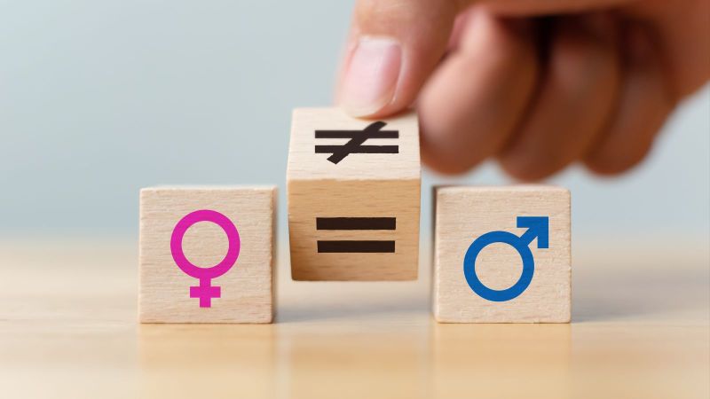 Das Bild zeigt drei Holzwürfel. Auf dem linken ist ein Weiblichsymbol, auf dem rechten ein Männlichkeitsymbol zu sehen. Der mittlere Würfel wird von einem Ungleichzeichen auf ein Gleichheitszeichen gedreht.