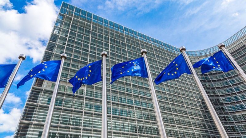 6 EU-Flaggen an Fahnenmasten vor einem mehrstöckigen Glasbau.