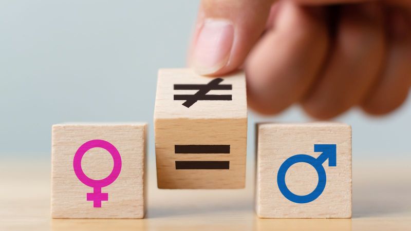 Das Bild zeigt drei Holzwürfel. Auf dem linken ist ein Weiblichsymbol, auf dem rechten ein Männlichkeitsymbol zu sehen. Der mittlere Würfel wird von einem Ungleichzeichen auf ein Gleichheitszeichen gedreht.
