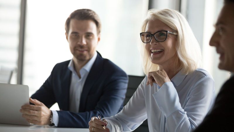 Das Bild zeigt eine Besprechung, bei der 3 Personen zu sehen sind. Eine Frau ist im Fokus gezeigt ist und an ihrer Seite sitzen zwei Herren.