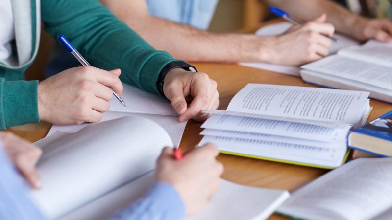 Drei Personen sitzen an einem Tisch und lesen in Büchern und schreiben Notizen dazu 