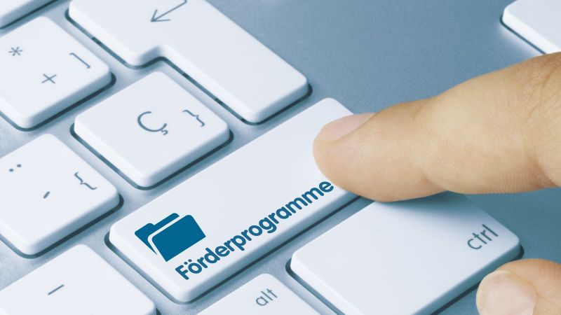 Das Bild zeigt eine Laptoptastatur auf der ein Finger eine Taste drückt, die als 'Förderprogramme' beschriftet ist.