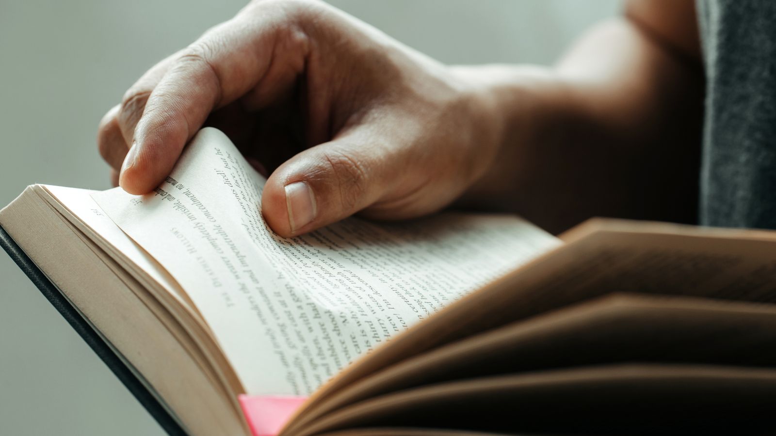 Eine Person liest in einem Buch. Zu sehen ist neben dem Buch mit lila Seitenmarkierung auch die rechte Hand, die gerade umblättert.
