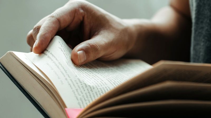 Eine Person liest in einem Buch. Zu sehen ist neben dem Buch mit lila Seitenmarkierung auch die rechte Hand, die gerade umblättert.