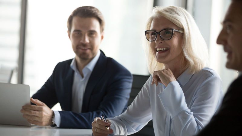 Das Bild zeigt eine Besprechung, bei der 3 Personen zu sehen sind. Eine Frau ist im Fokus gezeigt ist und an ihrer Seite sitzen zwei Herren.