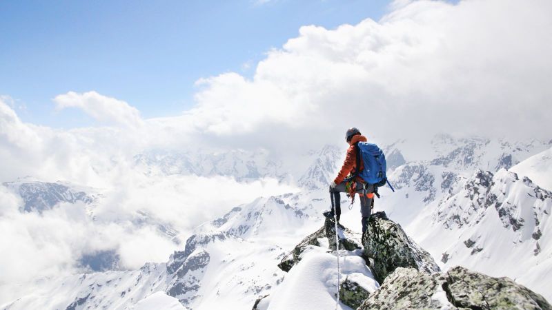 Eine bergsteigende Person ist auf einer Bergspitze zu sehen. Im Hintergrund sind schneebedeckte Berge zu sehen.