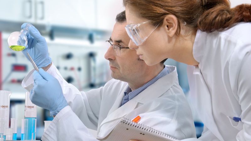 Das Bild zeigt einen Wissenschaftler und eine Wissenschaftler im Labor, die sich einen Rundkolben mit einer grünen Flüssigkeit ansehen 