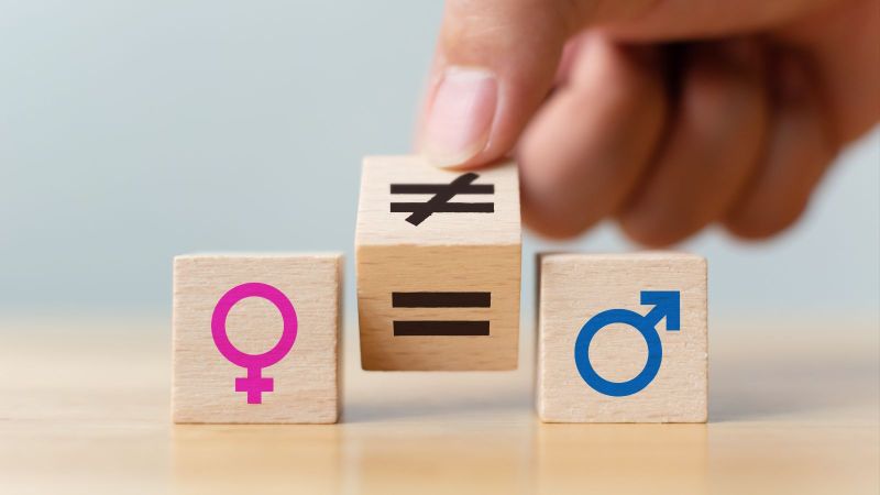 Das Bild zeigt drei Holzwürfel. Auf dem linken ist ein Weiblichsymbol, auf dem rechten ein Männlichkeitsymbol zu sehen. Der mittlere Würfel wird von einem Ungleichzeichen auf ein Gleichheitszeichen gedreht.