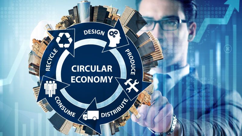 Das Bild zeigt einen Mann, der mit seiner Hand auf eine Zeichnung einer Stadt zeigt. Davor befindet sich ein blauer Kreislauf mit dem Wort Circular Economy im Zentrum und verschiedenen Begriffen die damit im Zusammenhang stehen.