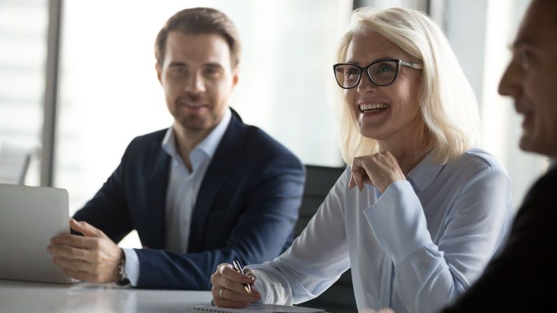 Das Bild zeigt eine Besprechung, bei der 3 Personen zu sehen sind. Eine Frau ist im Fokus gezeigt ist und an ihrer Seite sitzen zwei Herren.